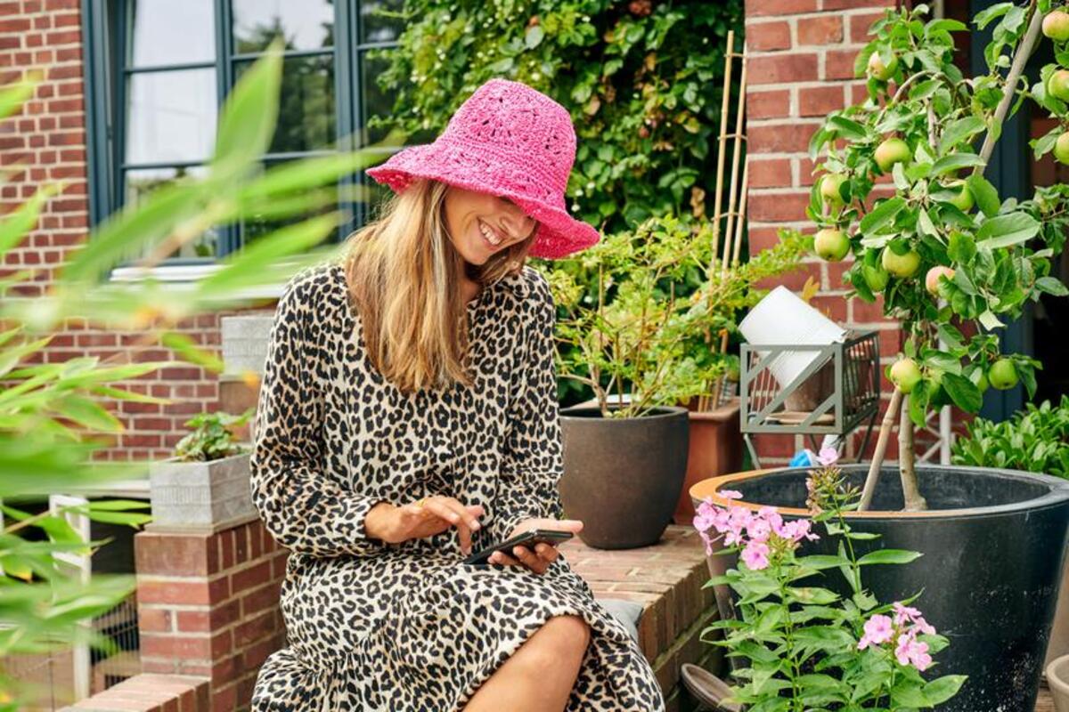 Uśmiechnięta kobieta w różowym kapeluszu siedzi przed domem i korzysta ze smartfona.
