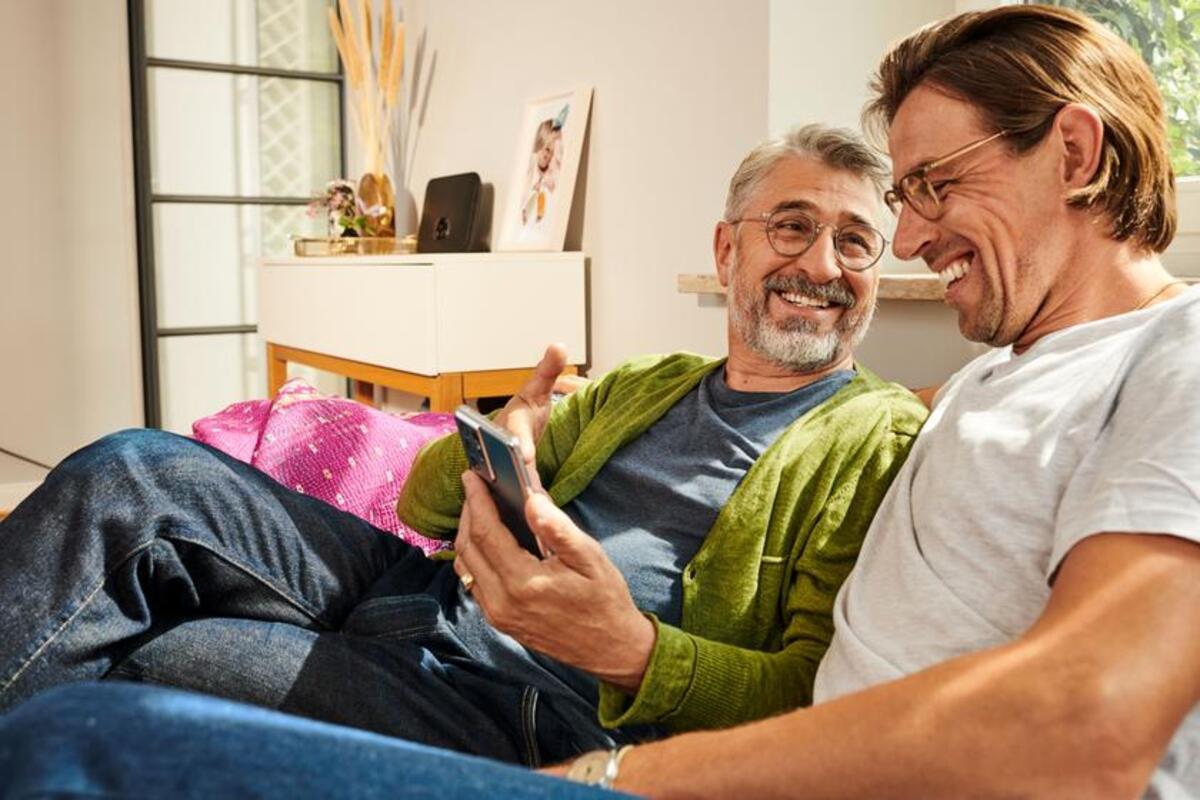 Uśmiechnięty starszy mężczyzna wskazuje na smartfon i patrzy na drugiego zadowolonego mężczyznę.