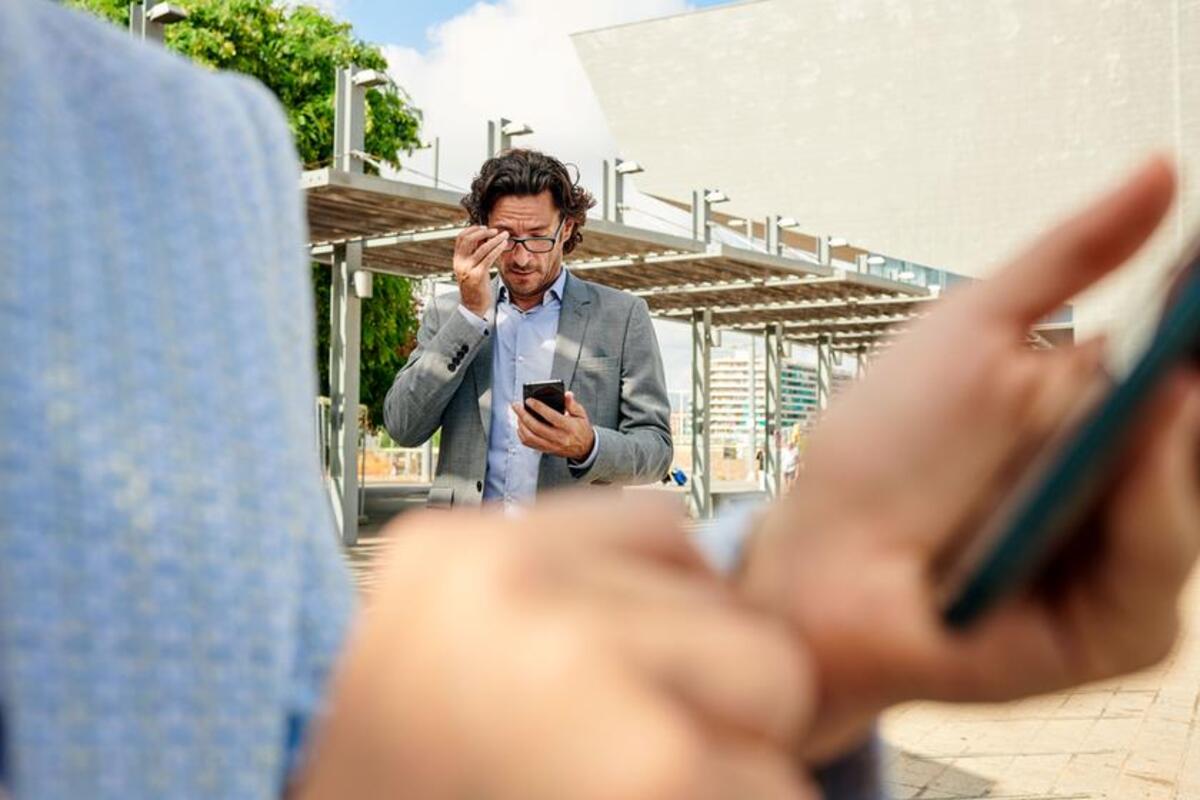 Na pierwszym planie rozmazana postać trzymająca telefon. W tle zaniepokojony mężczyzna parzący na telefon.